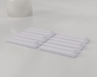 May include: A set of six white plastic rectangular components, each featuring a flat base with a small vertical tab at one end. These modular pieces are arranged in two parallel rows of three, showcasing a clean, uniform design.