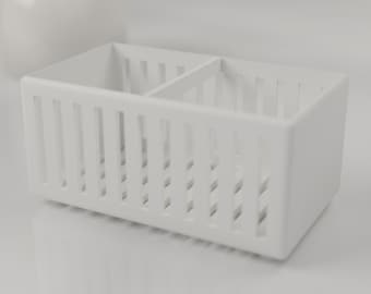 May include: A rectangular white organizer bin featuring a slatted design on all four sides and a central divider that splits the interior into two equal compartments. The structure is made of solid white material with vertical cutouts.