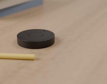 May include: A matte black circular smart button device with a centered circular press area and a small indicator light, positioned next to a thin yellow cylindrical object on a smooth, light-colored surface.