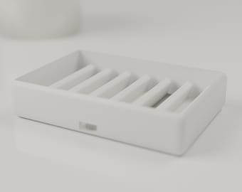 May include: A rectangular white soap dish featuring a series of parallel raised ridges along the interior base to allow for drainage. The exterior frame is smooth and solid with a small rectangular notch centered on the front side.