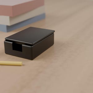 May include: A matte black rectangular note holder box with a lid and a front cutout, accompanied by a small yellow pencil, positioned near a stack of pastel pink, white, and blue paper sheets on a smooth surface.