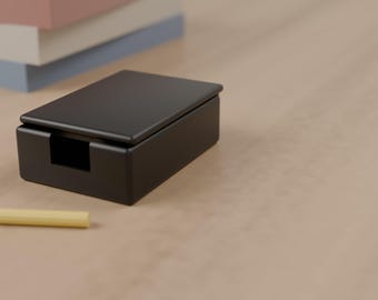 May include: A matte black rectangular note holder box with a lid and a front cutout, accompanied by a small yellow pencil, positioned near a stack of pastel pink, white, and blue paper sheets on a smooth surface.