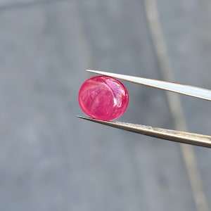 May include: A vibrant, round, cabochon-cut gemstone with a deep pinkish-red hue. The smooth, polished surface of the stone reflects light, showing internal clarity and colour saturation. The gemstone is held securely by a pair of silver-toned metal tweezers.