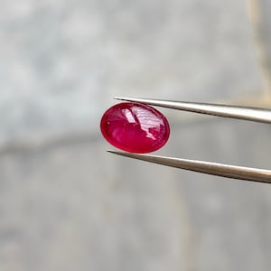 May include: A vibrant, oval-shaped, deep red gemstone held securely by a pair of silver-toned metal tweezers. The stone features a smooth, polished cabochon surface with visible internal inclusions and light reflections across its rounded top.