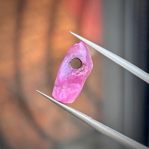 Op de afbeelding: Een ruwe, felroze edelsteen kraal met een onregelmatige, gefacetteerde oppervlaktestructuur en een enkel rond gat in het bovenste gedeelte. De steen wordt vastgehouden door een zilverkleurige metalen pincet, wat de doorschijnende en kristallijne structuur benadrukt.