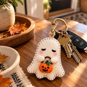 May include: A white felt ghost keychain featuring stitched edges, closed eyes, pink cheeks, and small arms holding a miniature orange jack-o'-lantern with a green stem. The ghost is attached to a gold-toned metal keyring with keys.