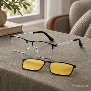Puede incluir: Gafas rectangulares con montura negra y plateada y lentes transparentes, acompañadas de un accesorio clip-on desmontable con lentes tintadas en amarillo. La montura tiene varillas plateadas y puntas negras. El texto VelloraEyewear es visible.