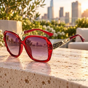Puede incluir: Gafas de sol rojas de gran tamaño con montura facetada y lentes degradadas. Las patillas presentan detalles en metal dorado con una incrustación negra brillante y texturizada, además de un diseño geométrico recortado. El texto VelloraEyewear es visible.