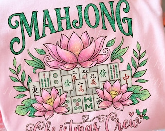 May include: A pale pink t-shirt featuring the word "MAHJONG" in green script, a lotus flower, and Mahjong tiles. The words "Christmas Crew" are written in a cursive font below. The design incorporates green leaves and pink flowers.