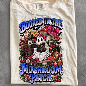 Può includere: Una t-shirt color crema con una stampa grafica che riporta il testo "BOOKED IN THE MUSHROOM PATCH". Il design include un fantasma che legge un libro, funghi, ciliegie, una candela, una lanterna e pipistrelli in un'illustrazione colorata.