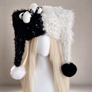 May include: A split-colour black and white fuzzy hat with long dangling sides ending in pom-poms. The black side features a white plush bone accessory secured with a black studded strap. The hat is displayed on a mannequin head with blonde hair.