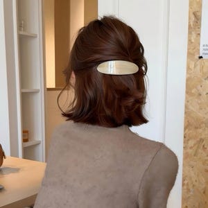 May include: A smooth, oval-shaped hair clip in a glossy cream color is fastened to dark brown hair. The hair clip has a simple, elongated design with a reflective surface, securing a section of hair at the back of the head.