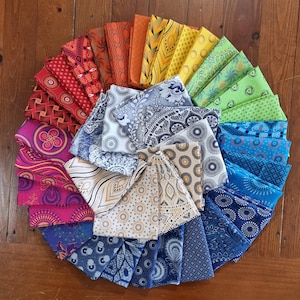 May include: A circular arrangement of folded fabric squares featuring a diverse spectrum of colours including red, orange, yellow, green, blue, and purple. Each square displays a unique pattern, such as geometric shapes, floral motifs, and dots.