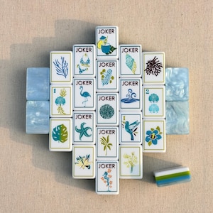 Könnte beinhalten: Eine Sammlung rechteckiger Mahjong-Spielsteine mit weißen Oberflächen, verziert mit blauen, grünen und orangefarbenen Illustrationen tropischer Flora, Fauna und Joker-Beschriftungen. Zwei hellblaue Marmorfliesen und eine gestreifte Fliese sind enthalten.