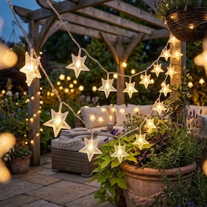 May include: A string of LED lights featuring clear, star-shaped covers. The lights emit a warm glow and are suspended across a wooden structure. Each star has a textured surface, and the strand is connected by a thin, flexible wire.