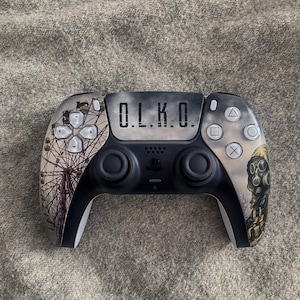 May include: A black and white wireless game controller featuring a custom graphic design. The center touchpad displays the text O.L.K.O. The left side shows a Ferris wheel illustration, while the right side depicts a figure wearing a gas mask.