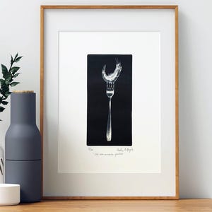 May include: A framed print featuring a black and white illustration of a fork with a shrimp resting on its tines. The artwork is set against a solid black rectangular background. Below the image, handwritten text reads "1/50" and "Claudio Pellegrini" with the phrase "Che bel momento gustoso!"