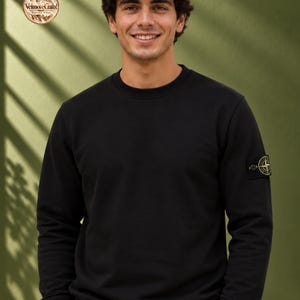 May include: A black long-sleeved crewneck sweatshirt featuring a circular embroidered patch on the left sleeve. The patch displays a yellow and black compass-style logo. The sweatshirt has ribbed cuffs and a ribbed hemline.