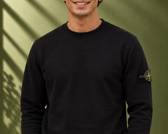 Op de afbeelding: Een zwart sweatshirt met lange mouwen en een ronde hals, voorzien van een ronde geborduurde patch op de linkermouw. De patch toont een geel en zwart logo in kompasstijl. Het sweatshirt heeft geribbelde boorden en een geribbelde zoom.