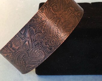 May include: A copper cuff bracelet with an intricate paisley pattern etched into the surface.