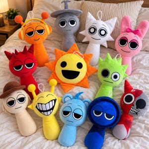 May include: A collection of twelve colorful plush toys featuring various character designs, including a sun, a skull-faced figure, and several creatures with antennae, headphones, or hats, all arranged together on a white fabric surface.