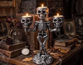 Puede incluir: Un candelabro de esqueleto en tono plateado con tres portavelas en forma de calavera. La figura central del esqueleto se apoya sobre una base decorada con calaveras adicionales, sosteniendo dos calaveras en sus manos, cada una con una vela encendida.
