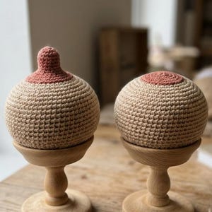Puede incluir: Dos objetos de ganchillo con forma de pecho hechos de hilo beige con detalles de areola y pezón en color marrón rosado. Cada pieza está montada sobre un soporte de madera clara con base redonda y tallo torneado.