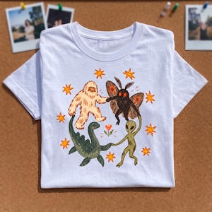 Può includere: Una t-shirt bianca a maniche corte con una stampa grafica colorata raffigurante un Bigfoot, una creatura simile a una falena, un mostro marino verde e un alieno che si tengono per mano in cerchio, circondati da piccole stelle arancioni.