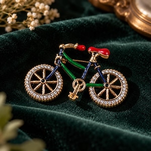 May include: A bicycle-shaped brooch featuring a gold-tone metal frame with green and dark blue enamel accents. The wheels are lined with small clear crystals, and the seat and handlebars are finished with a bright red enamel coating.