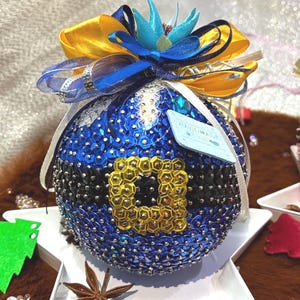 Pode incluir: Um ornamento esférico azul coberto de lantejoulas com um cinto de lantejoulas pretas e uma fivela de lantejoulas douradas. O topo é decorado com um laço de fita multicolorido em azul, amarelo e branco. Uma pequena etiqueta está presa ao lado.