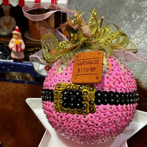 Pode incluir: Um enfeite de Natal esférico coberto de lantejoulas cor-de-rosa com um cinto de lantejoulas pretas e uma fivela dourada. Uma etiqueta de couro castanho diz HANDMADE WITH LOVE. O enfeite é rematado com um laço dourado e uma fita rosa.