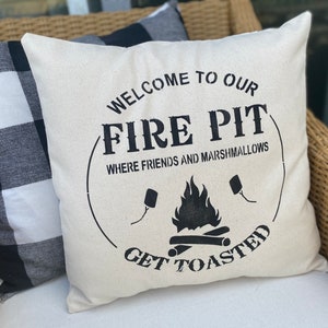 May include: White throw pillow with black text that reads "Welcome to our Fire Pit Where Friends and Marshmallows Get Toasted". The pillow has a black and white checkered pillow behind it.