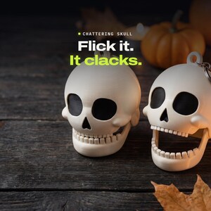 May include: Two off-white skull keychains with black eye sockets and nasal cavities. The text CHATTERING SKULL Flick it. It clacks. is printed above the skulls. One skull has a closed jaw, while the other has an open jaw showing teeth.
