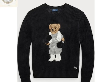 Könnte beinhalten: Ein schwarzer Langarmpullover mit Rundhalsausschnitt und strukturiertem Strickmuster. In der Mitte befindet sich eine gestickte Grafik eines braunen Bären, der ein graues Sweatshirt mit dem Text POLO, graue Shorts und weiße Stiefel trägt. Die Buchstaben RL sind unten rechts in Weiß gestickt.