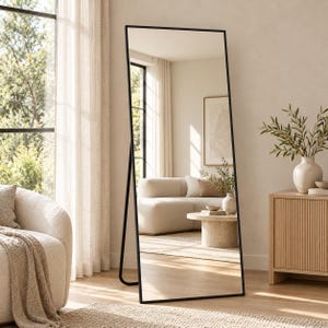 May include: A tall, rectangular floor mirror featuring a thin, black metal frame. The mirror is designed to stand independently, supported by a matching black metal stand attached to the back, reflecting a bright, neutral-toned living room.