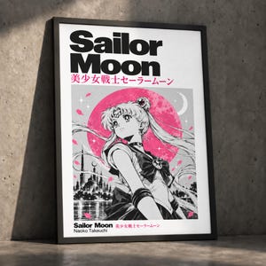 Op de afbeelding: Een ingelijste poster met de tekst Sailor Moon en Japanse karakters. Het kunstwerk toont een zwart-wit illustratie van het personage Sailor Moon tegen een grote roze maan, met een stadssilhouet onderaan en zwevende roze bloemblaadjes.