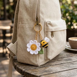 Puede incluir: Una mochila de lona beige tiene un llavero de metal dorado sujeto a la cremallera. El llavero consta de una margarita blanca de ganchillo con el centro amarillo y una pequeña hoja verde, junto a una abeja de ganchillo a rayas amarillas y negras.
