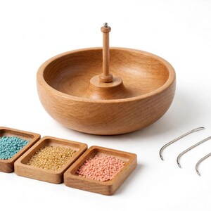 May include: A wooden bead spinner bowl with a central vertical spindle, three small rectangular wooden trays containing turquoise, gold, and pink seed beads, and three curved metal beading needles on a plain white background.