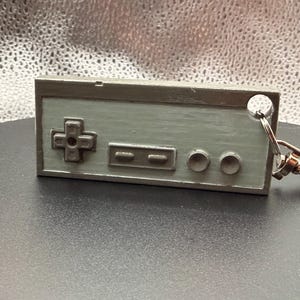 May include: A gray, rectangular keychain resembling a classic video game controller. It features a directional pad, two rectangular buttons, and two round buttons. A silver keyring and clasp are attached.