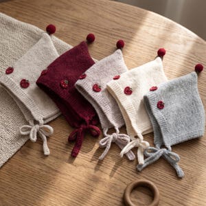 Puede incluir: Un conjunto de cinco gorros tejidos en beige, burdeos, rosa claro, crema y gris. Cada gorro presenta una parte superior puntiaguda con un pequeño pompón rojo, dos pequeñas aplicaciones de fresa roja en el lateral y lazos a juego.