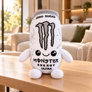 May include: A white plush toy shaped like a Monster Energy Ultra can with a grey top, two arms, and two feet. The front features the black Monster logo, the text ZERO SUGAR, MONSTER ENERGY ULTRA, and a smiling face with two large black eyes.