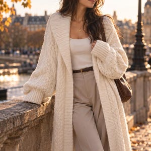 Könnte beinhalten: Eine lange, cremefarbene Strickjacke mit Zopfmuster, breitem Schalkragen und gerippten Bündchen, getragen über einem weißen Tanktop und einer beigen Hose mit einem dünnen braunen Ledergürtel. Eine braune Lederschultertasche wird seitlich getragen.