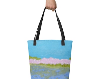 Puede incluir: Un bolso tote con un paisaje impreso que muestra un cielo azul brillante, una hilera de árboles con flores de color rosa claro y un campo texturizado en tonos azules y verdes. El bolso tiene dos asas de tela negra en la parte superior.