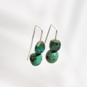 Puede incluir: Pendientes colgantes con dos piedras redondeadas verde-azuladas suspendidas de ganchos plateados. Las piedras tienen un aspecto moteado con acentos verdes oscuros y marrones, creando una estética natural y terrosa. Sobre un fondo blanco liso.