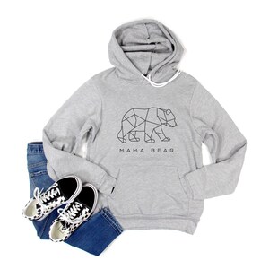May include: A light gray hoodie with a geometric bear design and the text "MAMA BEAR". The hoodie is paired with blue jeans and black and white patterned sneakers. The hoodie has a front pocket and a drawstring hood.