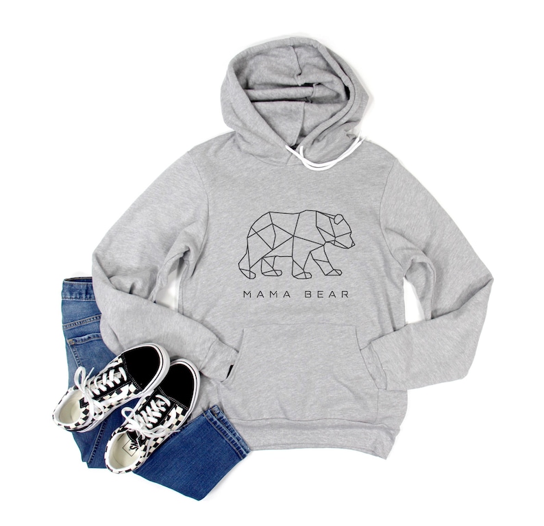 May include: A light gray hoodie with a geometric bear design and the text "MAMA BEAR". The hoodie is paired with blue jeans and black and white patterned sneakers. The hoodie has a front pocket and a drawstring hood.