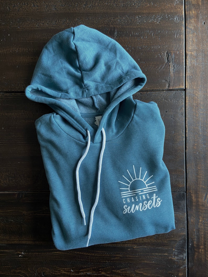 Puede incluir: Una sudadera con capucha azul turquesa con cordones blancos y un gr&aacute;fico blanco de una puesta de sol con el texto "Chasing sunsets" en la parte delantera.
