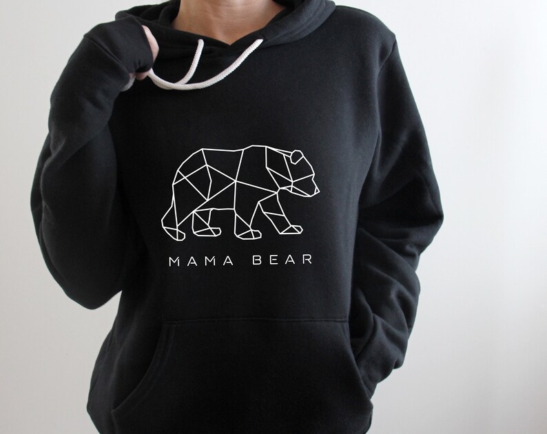 May include: Black hoodie with a white geometric bear design and the text "MAMA BEAR". The hoodie has a drawstring hood and a front pocket. The bear graphic is a modern, minimalist design.