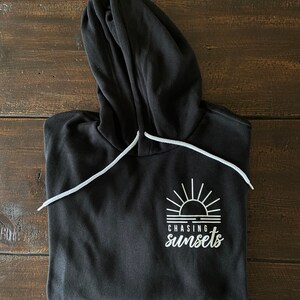 Puede incluir: Sudadera con capucha negra con un gr&aacute;fico blanco de un sol poni&eacute;ndose sobre el agua y el texto "CHASING sunsets".