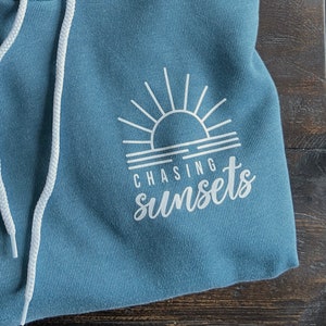 Puede incluir: Una sudadera con capucha azul con un gr&aacute;fico blanco de una puesta de sol y el texto "CHASING sunsets".